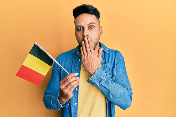 Молодий арабський чоловік тримає прапор бельгії, покриваючи рот рукою, шокований і боїться помилки. здивований вираз
  - Фото, зображення