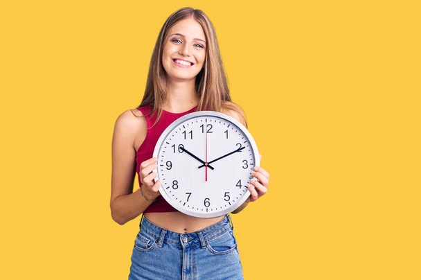 Young beautiful blonde woman holding big clock looking positive and happy standing and smiling with a confident smile showing teeth  - Photo, image