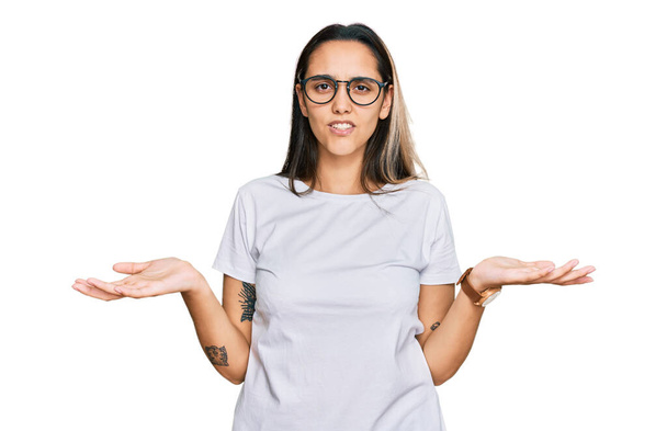 Jonge Latijns-Amerikaanse vrouw draagt casual wit t-shirt onwetend en verward met open armen, geen idee concept.  - Foto, afbeelding