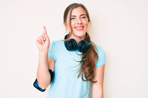 Schöne junge kaukasische Frau in Turnbekleidung und mit Kopfhörern zeigt und zeigt mit Finger Nummer eins, während sie selbstbewusst und glücklich lächelt.  - Foto, Bild