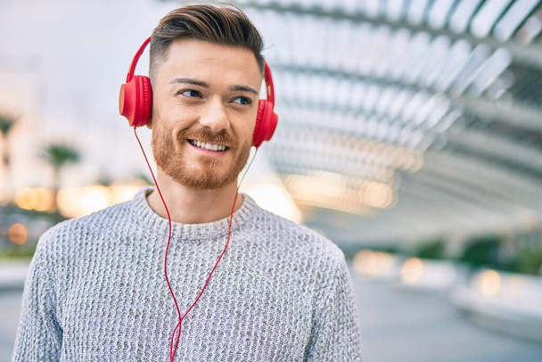 Νεαρός καυκάσιος άνδρας χαμογελά χαρούμενος ακούγοντας μουσική χρησιμοποιώντας ακουστικά στην πόλη. - Φωτογραφία, εικόνα