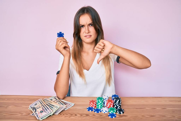 Красивая белая женщина, играющая в покер, держит фишки казино с сердитым лицом, негативный знак, показывающий неприязнь с опущенными пальцами, концепция отказа  - Фото, изображение