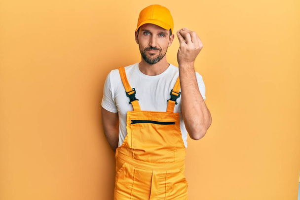 Jonge knappe man draagt klusjesman uniform over gele achtergrond doet italiaans gebaar met de hand en vingers zelfverzekerde uitdrukking  - Foto, afbeelding