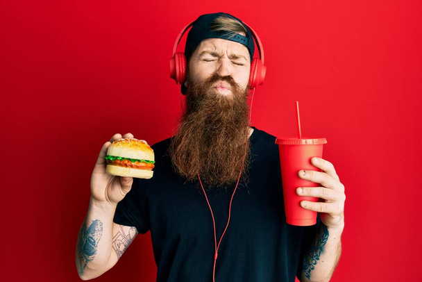 Rudowłosy mężczyzna z długą brodą jedzący smacznego, klasycznego burgera i pijącego sodę patrzącego w obiektyw, dmuchający pocałunek, jest uroczy i seksowny. wyrażenie miłości.  - Zdjęcie, obraz