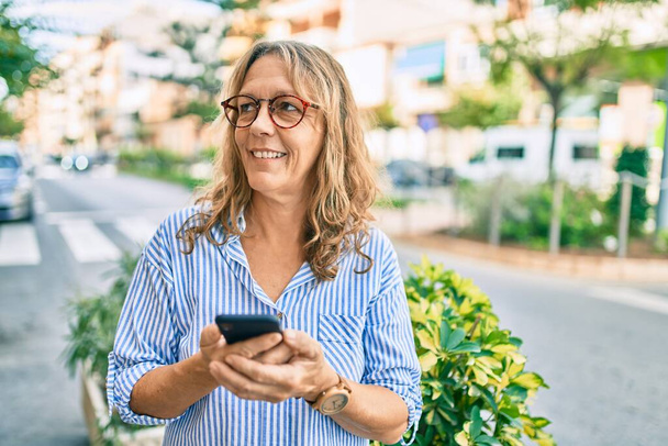 Mulher caucasiana de meia idade sorrindo feliz usando smartphone na cidade. - Foto, Imagem