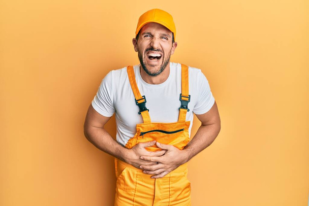 Young handsome man wearing handyman uniform over yellow background smiling and laughing hard out loud because funny crazy joke with hands on body.  - Фото, зображення