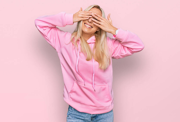 Young blonde woman wearing casual sweatshirt covering eyes with hands smiling cheerful and funny. blind concept.  - Фото, изображение