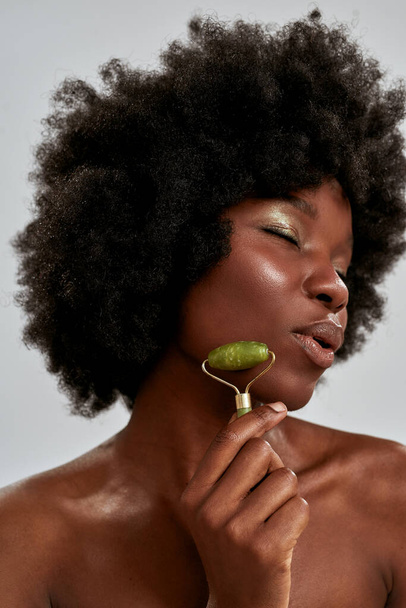 Close up portrait of attractive young african american woman with perfect glowing skin using jade roller or natural massage tool, posing isolated over gray background - Valokuva, kuva