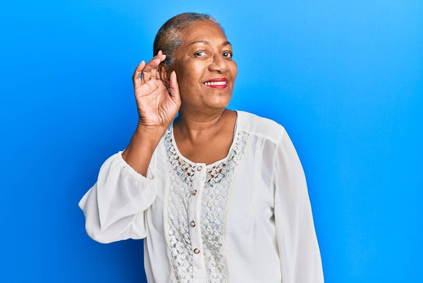 Mulher americana africana sênior vestindo roupas casuais sorrindo com a mão sobre orelha ouvindo e ouvindo rumores ou fofocas. conceito de surdez.  - Foto, Imagem