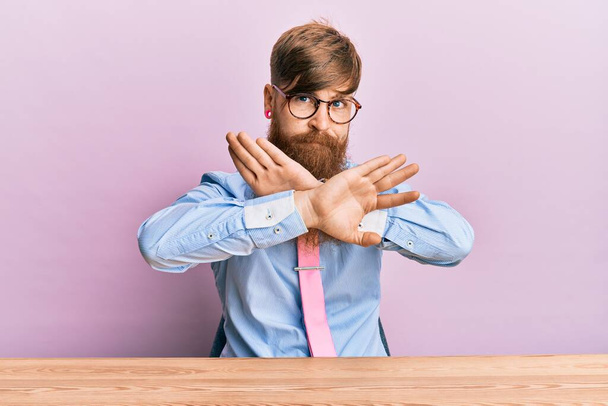 若いアイルランドの赤毛の男がビジネスシャツを着てネクタイをテーブルの上に座って否定的な記号を行う腕や手のひらを横断拒否式怒りの顔  - 写真・画像