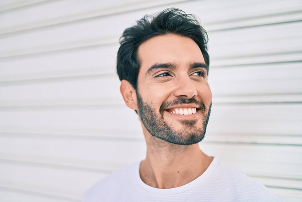 Joven hombre hispano sonriendo feliz apoyado en la pared de la ciudad. - Foto, imagen