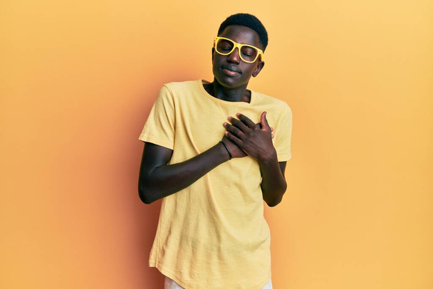 Young african american man wearing casual clothes and glasses smiling with hands on chest with closed eyes and grateful gesture on face. health concept.  - Fotografie, Obrázek