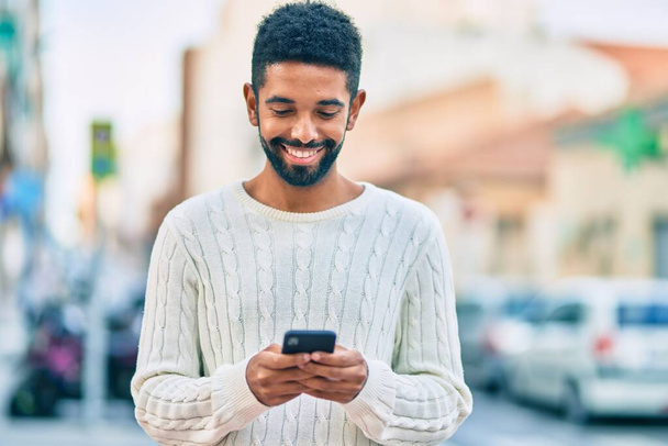 Jeune homme afro-américain souriant heureux en utilisant un smartphone à la ville. - Photo, image