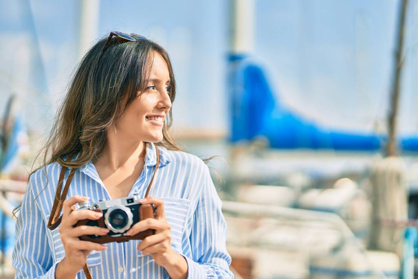 Young hispanic tourist woman smiling happy using vintage camera at the port. - Фото, изображение