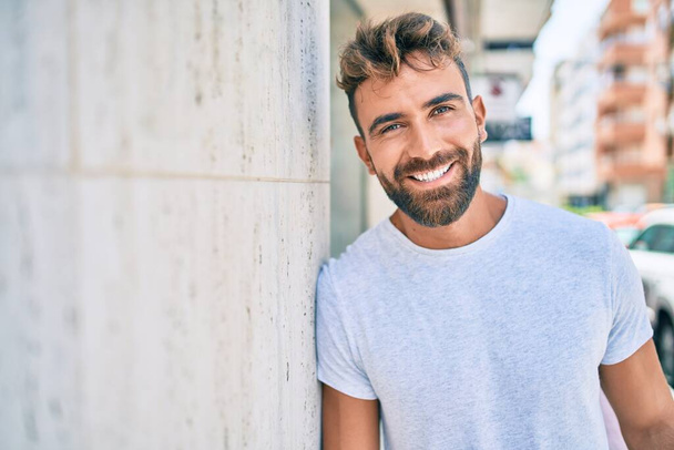 Fiatal spanyol férfi mosolyog boldogan támaszkodik a falra a városban. - Fotó, kép