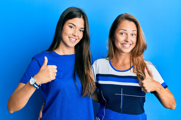 Spanyol család anya és lánya viselnek alkalmi ruhát kék háttér csinál boldog hüvelykujj fel gesztus kézzel. jóváhagyása kifejezés nézi a kamera mutatja siker.  - Fotó, kép