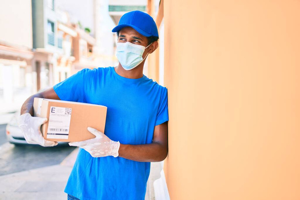 Africano homem de entrega vestindo correio uniforme ao ar livre usando máscara de segurança coronavírus segurando pacote de papelão - Foto, Imagem