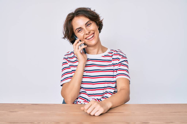 Fiatal spanyol nő beszél az okostelefonon ül az asztalon pozitív és boldog álló és mosolygó magabiztos mosollyal mutatja fogak  - Fotó, kép