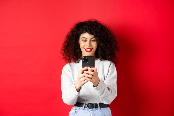 Souriant fille moderne prenant des photos sur smartphone, regardant l'écran et enregistrant la vidéo, debout sur fond rouge - Photo, image