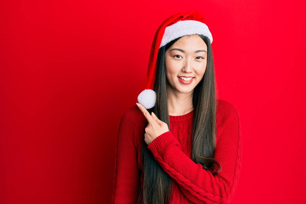 Genç Çinli kadın Noel şapkası takıyor. Gülümsüyor, gülümsüyor. Elleri ve parmakları yana bakıyor.  - Fotoğraf, Görsel