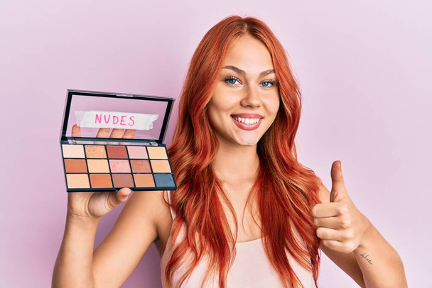 Young redhead woman holding makeup nudes smiling happy and positive, thumb up doing excellent and approval sign  - Photo, Image
