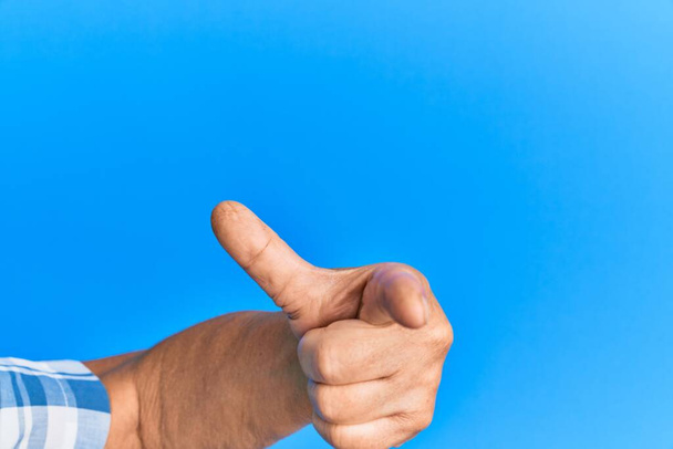 Mano del hombre hispano mayor sobre fondo azul aislado apuntando con el dedo índice a la cámara, eligiendo e indicando dirección  - Foto, Imagen