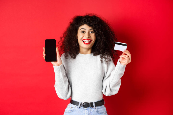 Elektronikus kereskedelem és online vásárlási koncepció. Vidám nő mosolyog, mutatja műanyag hitelkártya és üres okostelefon képernyő, álló piros háttér - Fotó, kép