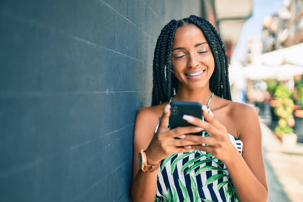 Junge Afroamerikanerin lächelt glücklich mit Smartphone in die Stadt. - Foto, Bild
