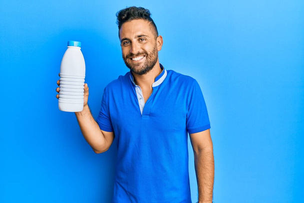 Handsome man with beard holding liter bottle of milk looking positive and happy standing and smiling with a confident smile showing teeth  - Photo, image