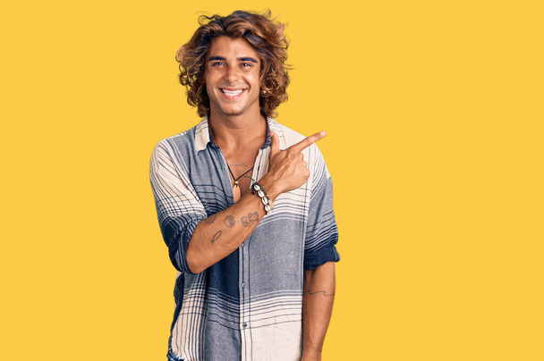 Молодой латиноамериканец в летнем стиле веселый с улыбкой на лице, указывающей рукой и пальцем в сторону со счастливым и естественным выражением лица  - Фото, изображение