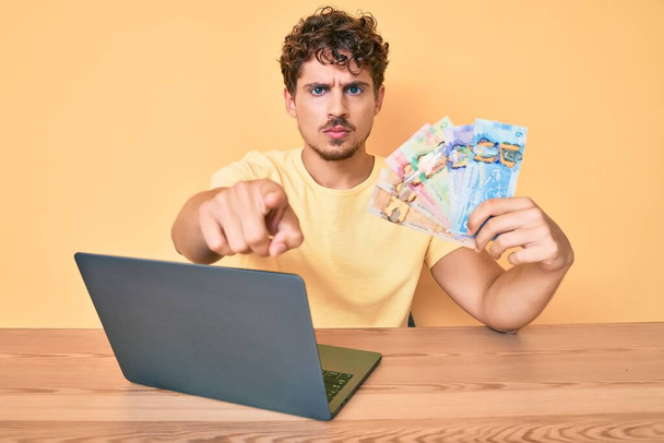 Молодий білий чоловік з кучерявим волоссям сидить на столі, працюючи з ноутбуком і тримає канадські банкноти доларів, вказуючи пальцем на камеру і до вас, впевнений жест виглядає серйозно
  - Фото, зображення