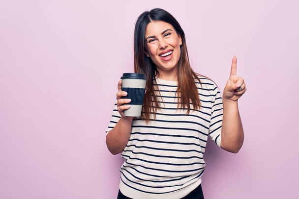 Молодая красивая брюнетка женщина пьет чашку кофе на вынос на изолированном розовом фоне улыбаясь с идеей или вопрос указывая пальцем со счастливым лицом, номер один - Фото, изображение