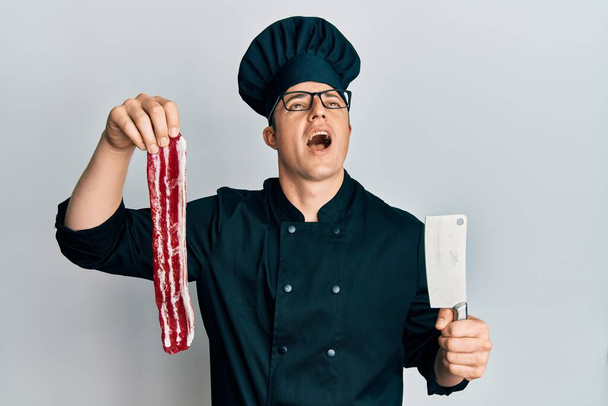 Handsome young man wearing chef uniform holding bacon and knife angry and mad screaming frustrated and furious, shouting with anger looking up.  - Photo, Image