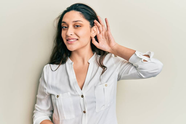 Bella donna mediorientale che indossa abiti casual sorridendo con mano sopra l'orecchio ascoltando una voce o pettegolezzi. concetto di sordità.  - Foto, immagini