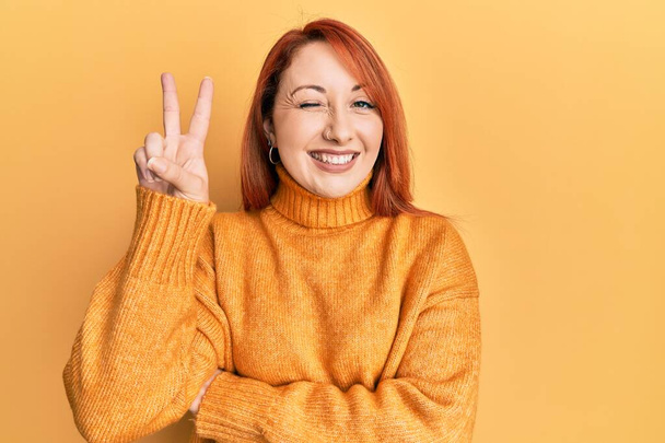 Beautiful redhead woman wearing casual winter sweater over yellow background smiling with happy face winking at the camera doing victory sign with fingers. number two.  - Photo, Image