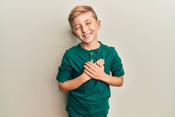 Kleiner kaukasischer Junge in lässiger Kleidung mit lächelnden Händen auf der Brust und geschlossenen Augen mit dankbarer Geste im Gesicht. Gesundheitskonzept.  - Foto, Bild