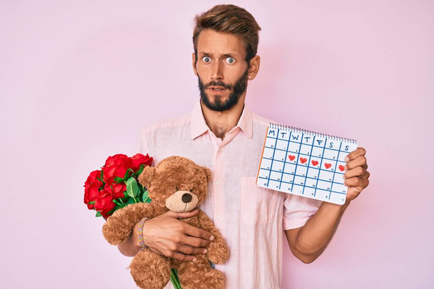 Handsome caucasian man with beard holding bear and bouquet of flowers for anniversary in shock face, looking skeptical and sarcastic, surprised with open mouth  - Photo, Image