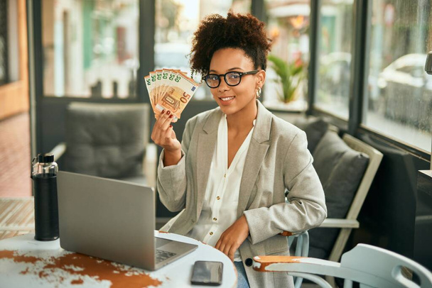 Young african american businesswoman holding 50 euro banknotes sitting at coffee shop terrace - Foto, Imagen