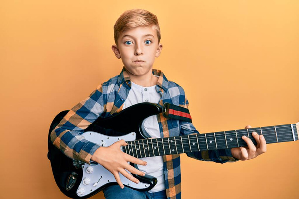 Kis kaukázusi srác elektromos gitáron játszik puffadó arccal, vicces arccal. levegővel felfújt száj, levegőbefúvás.  - Fotó, kép