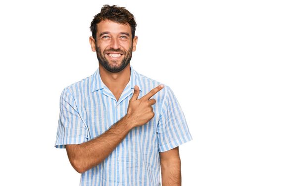 Όμορφος νεαρός άνδρας με γενειάδα φορώντας casual φρέσκο πουκάμισο χαρούμενο με χαμόγελο στο πρόσωπο δείχνοντας με το χέρι και το δάχτυλο στο πλάι με χαρούμενη και φυσική έκφραση στο πρόσωπο  - Φωτογραφία, εικόνα