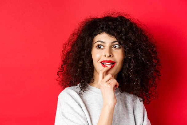 Close-up of excited curly woman touching lips and looking aside with interest, checking out cool promo, standing against red background - Foto, Imagem