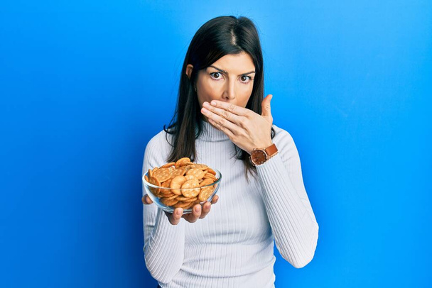 Mujer hispana joven sosteniendo galletas saladas cuenco cubriendo la boca con la mano, conmocionada y temerosa de equivocarse. expresión sorprendida  - Foto, Imagen