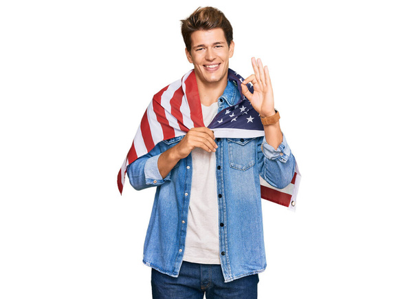 Hübscher kaukasischer Mann, der die Flagge der Vereinigten Staaten in der Hand hält und mit den Fingern Okay-Zeichen macht, freundlich lächelnd, hervorragendes Symbol gestikulierend  - Foto, Bild