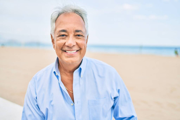 Handosme homme hispanique aux cheveux gris souriant heureux à la plage, profitant de vacances en été - Photo, image