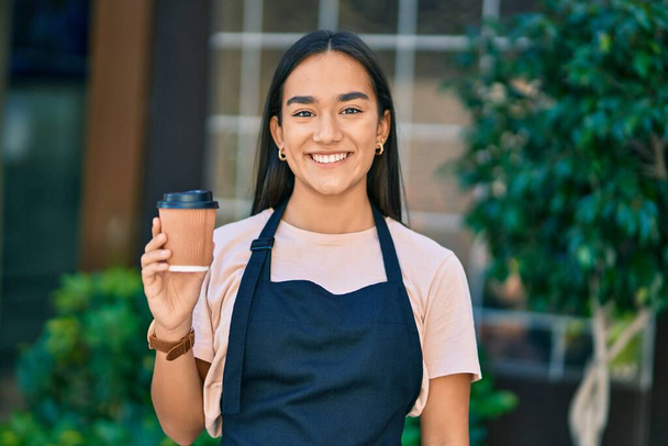 Young latin shopkeeper girl smiling happy drinking coffee at the city - Foto, Imagem