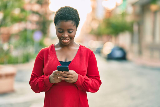 Junge Afroamerikanerin lächelt glücklich mit Smartphone in die Stadt. - Foto, Bild