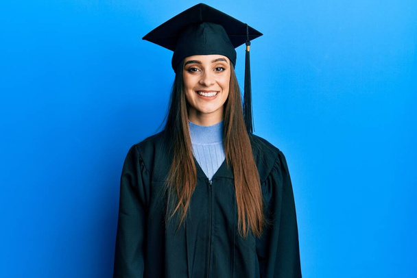 Belle jeune femme brune portant une casquette de graduation et une robe de cérémonie avec un sourire heureux et cool sur le visage. chanceux.  - Photo, image