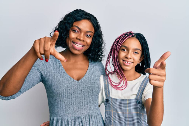 Bela mãe e filha afro-americana vestindo roupas casuais e abraçando dedos apontando para a câmera com rosto feliz e engraçado. boa energia e vibrações.  - Foto, Imagem