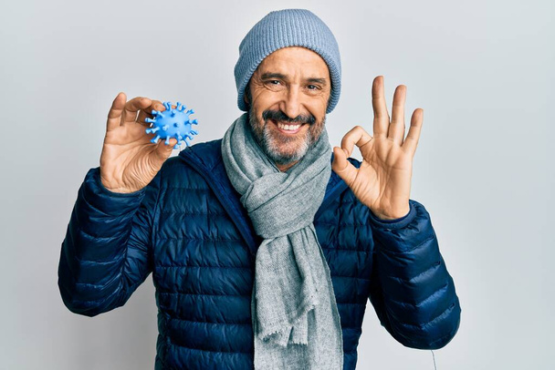 Middle age hispanic man wearing winter style holding virus toy doing ok sign with fingers, smiling friendly gesturing excellent symbol  - Photo, Image