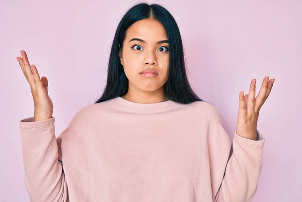 Young beautiful asian girl wearing casual pink sweater clueless and confused with open arms, no idea and doubtful face.  - Photo, Image